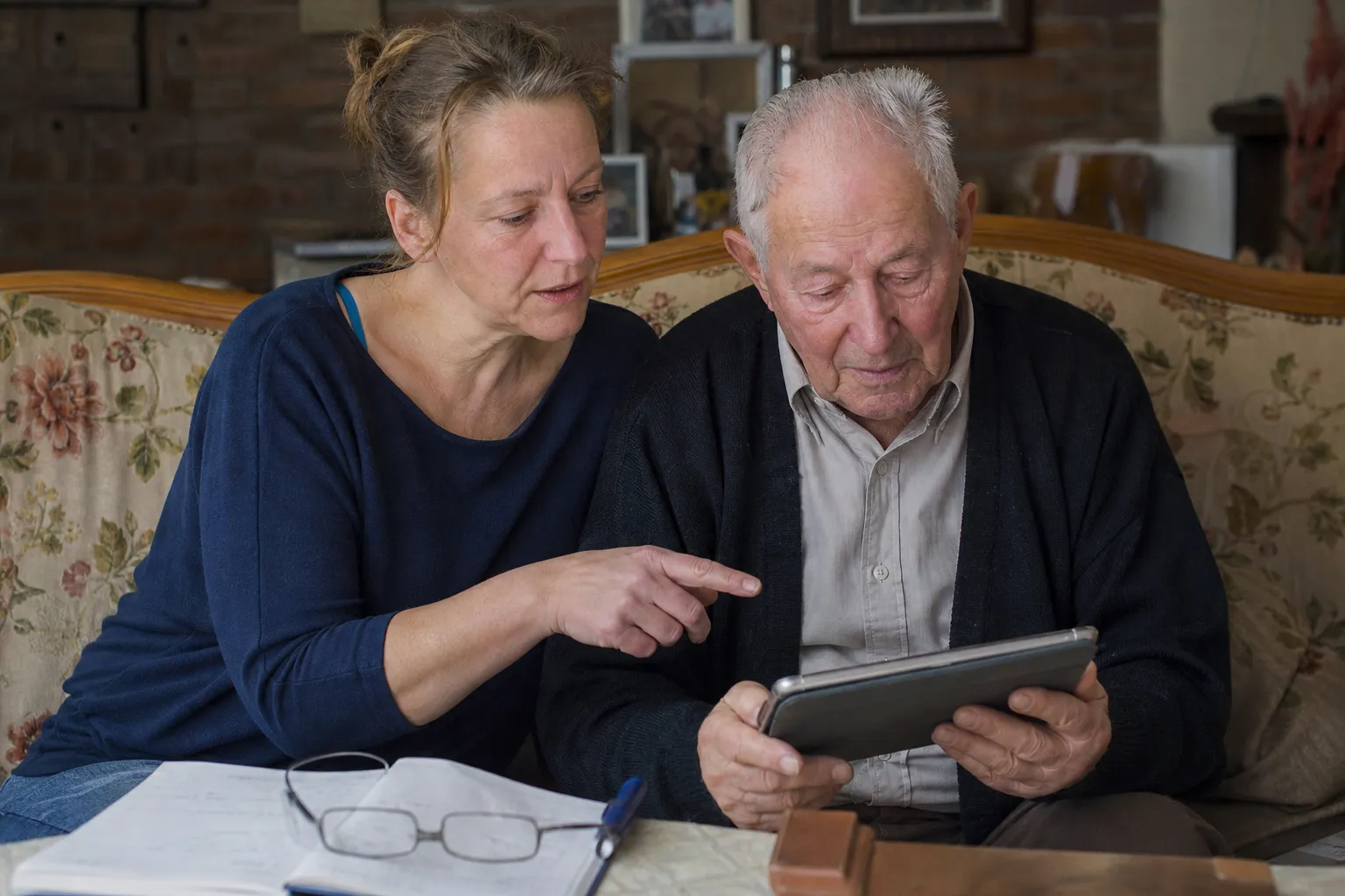 Une femme d'une quarantaine d'années montre une tablette à son père âgé, assis à côté d'elle dans son salon.