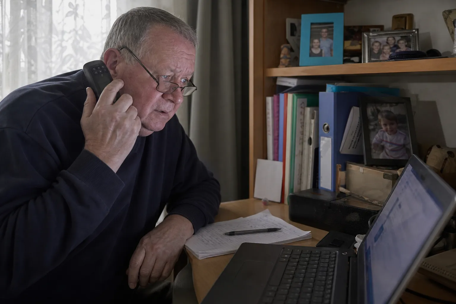 Un homme d'une soixantaine d'années, téléphone à l'oreille, regarde d'un air inquiet l'écran de son ordinateur portable.