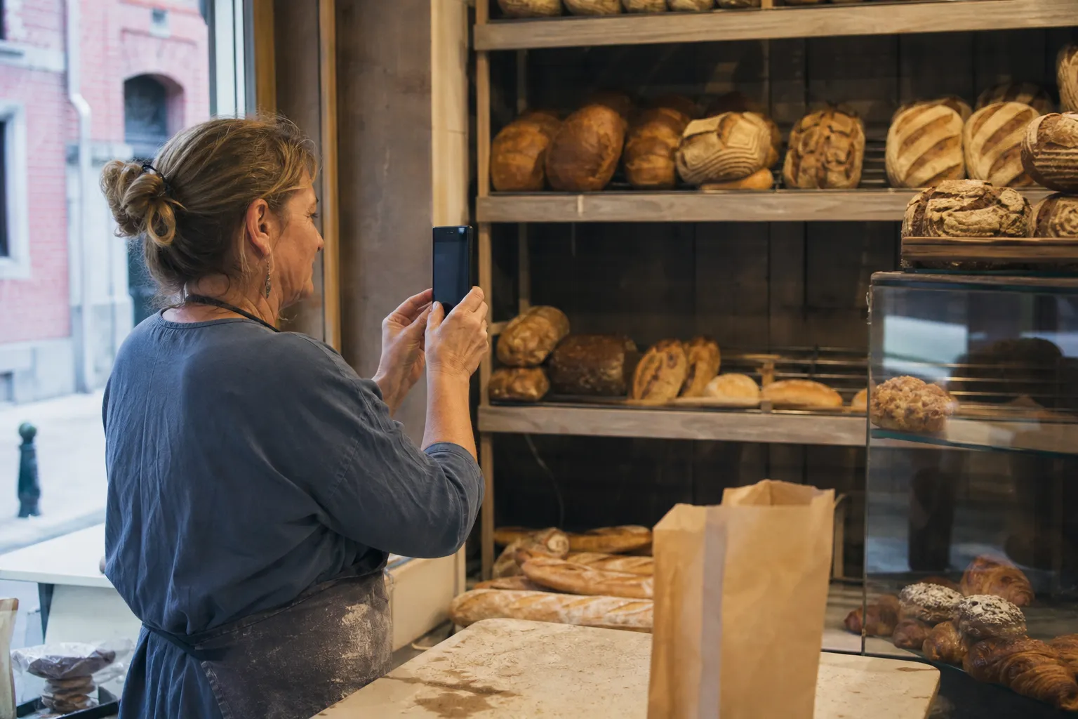 Une boulangère photographie avec son smartphone une vitrine de pains et de viennoiseries dans sa boutique.
