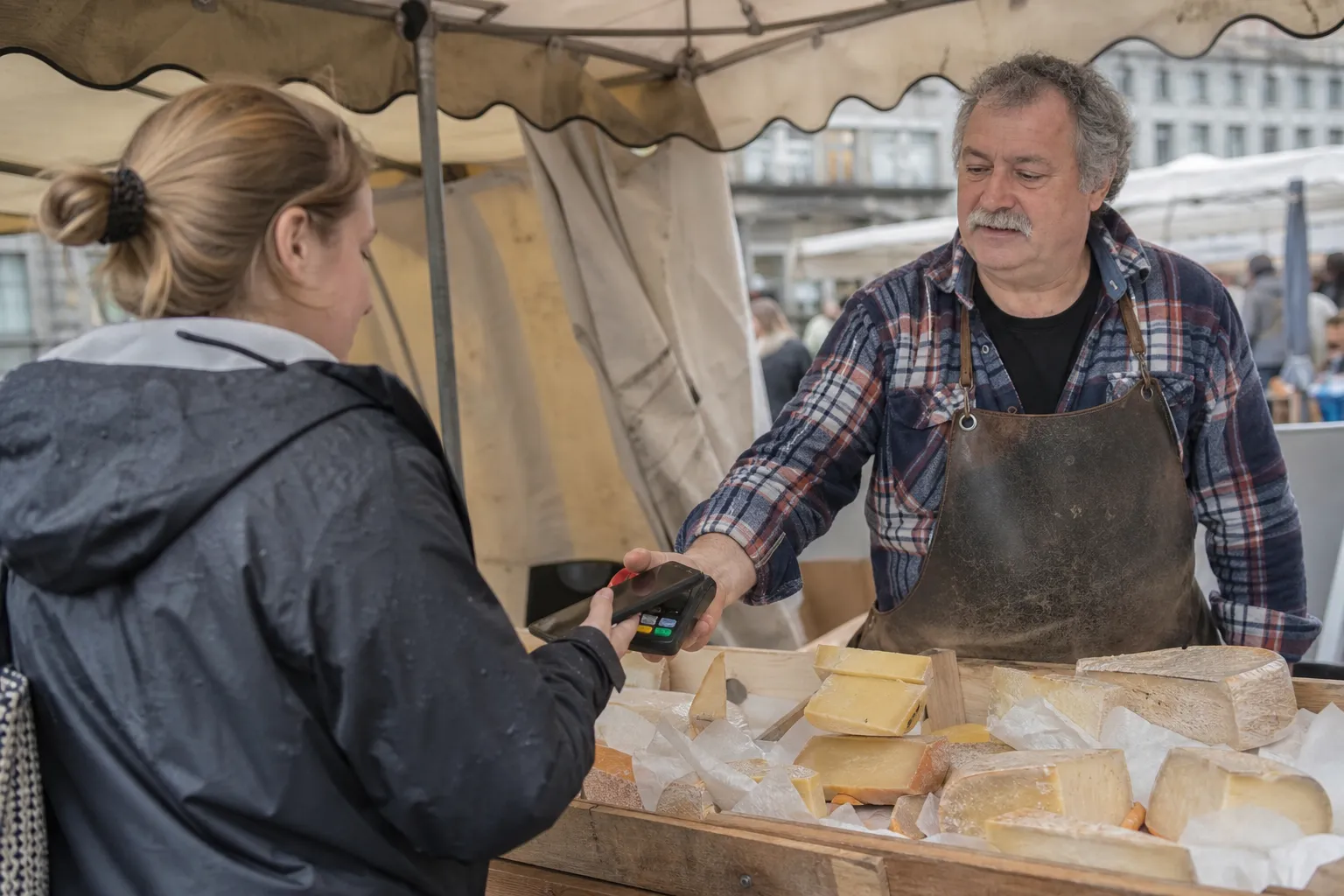 Un fromager tend un terminal de paiement portable à une cliente qui paie avec son smartphone sur un marché.