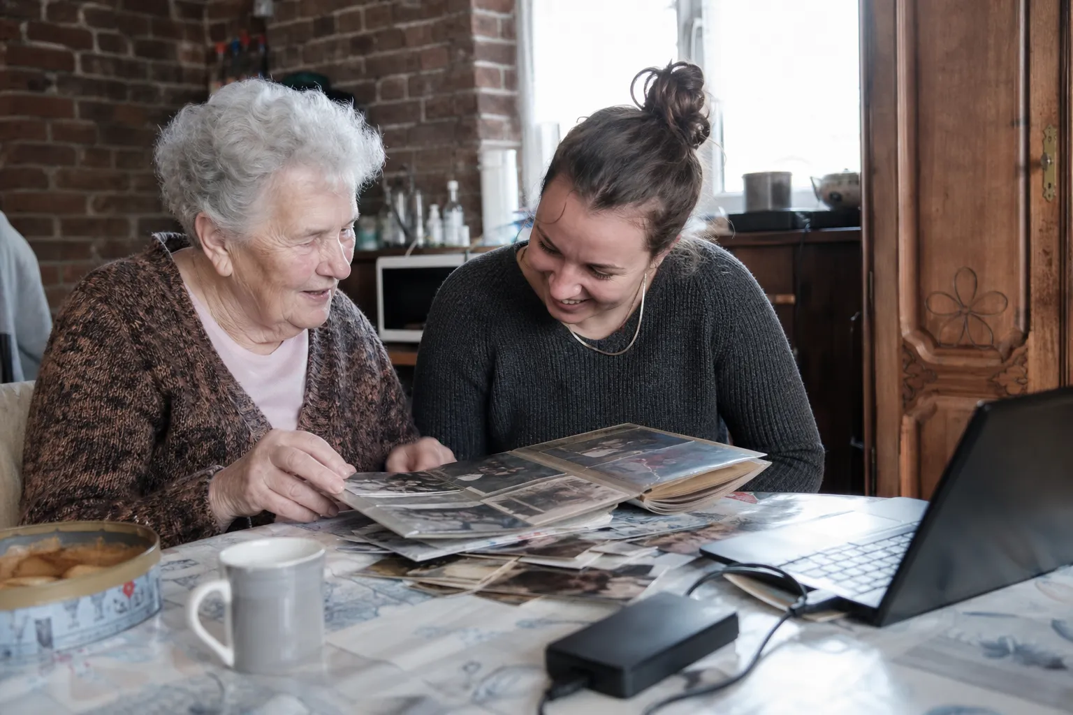 Une grand-mère et sa petite-fille trient de vieux albums photo étalés sur une table, un ordinateur portable ouvert à côté.