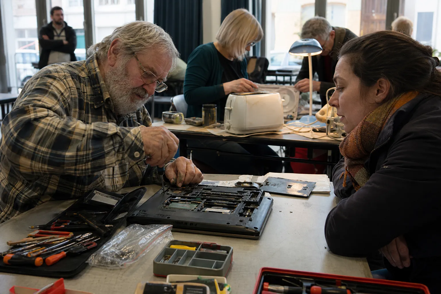 Un bénévole répare un ordinateur portable ouvert sur une table lors d'un Repair Café à Charleroi, sous le regard de sa propriétaire.