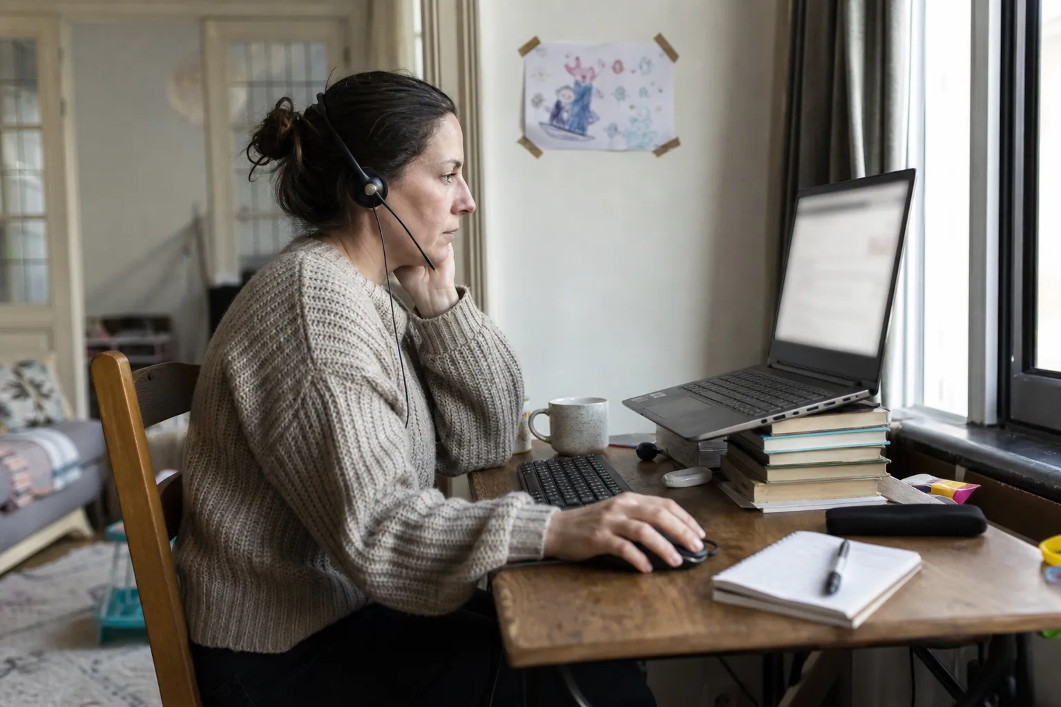 Une femme d'une trentaine d'années travaille sur un ordinateur portable surélevé, avec un casque, dans un coin bureau aménagé près d'une fenêtre.