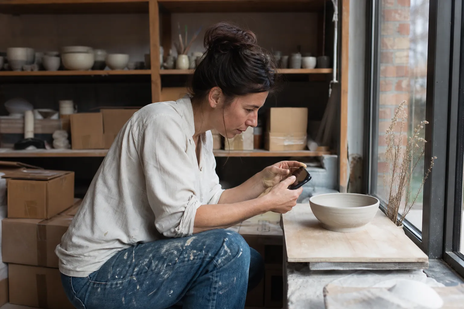 Une céramiste photographie un bol posé sur une planche près de la fenêtre de son atelier.